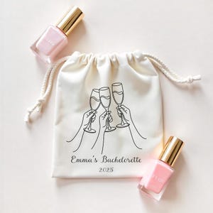 Op de afbeelding: Een witte katoenen trekkoordtas met een zwarte lijntekening van drie champagne glazen met de tekst "Emma's Bachelorette 2025" gedrukt op de voorkant.