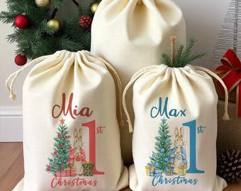 May include: Two personalized Christmas gift bags with the names Mia and Max, each with a number 1 and the words "1st Christmas". The bags are decorated with a Peter Rabbit-inspired design featuring a rabbit holding a gift and a Christmas tree.