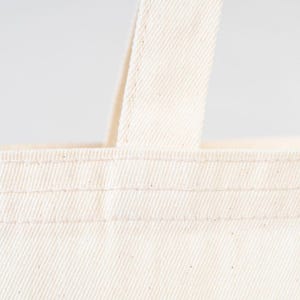 May include: Close-up of a white canvas tote bag with double stitched seams. The bag has a single handle.