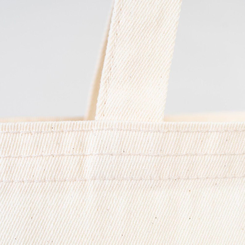 May include: Close-up of a white canvas tote bag with double stitched seams. The bag has a single handle.