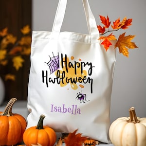 Puede incluir: Bolsa de tela blanca con el texto "Happy Halloween" en negro y morado. Una pequeña araña con un cuerpo morado y patas negras está debajo del texto. El nombre "Isabella" está impreso en morado debajo de la araña.