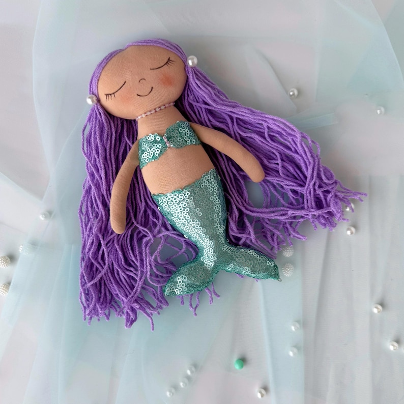 May include: A handmade purple haired mermaid doll with a teal sequin tail. The doll is lying on a light blue fabric background.