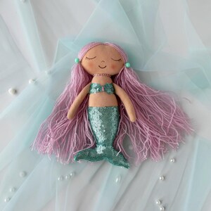 May include: A handmade mermaid doll with pink yarn hair, a green sequin tail, and a light pink seashell top. The doll is lying on a light blue tulle fabric.