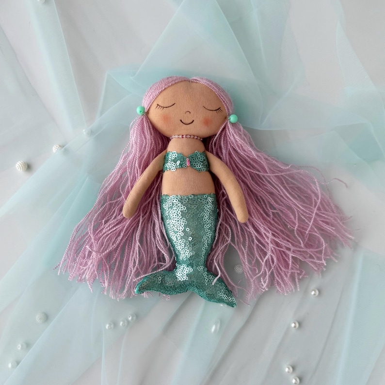 May include: A handmade mermaid doll with pink yarn hair, a green sequin tail, and a light pink seashell top. The doll is lying on a light blue tulle fabric.