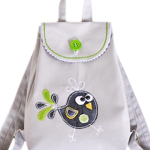 May include: A light gray backpack with a flap closure and a green button. The backpack features an embroidered cartoon bird with black, yellow, and green accents. The straps and handle are also light gray.