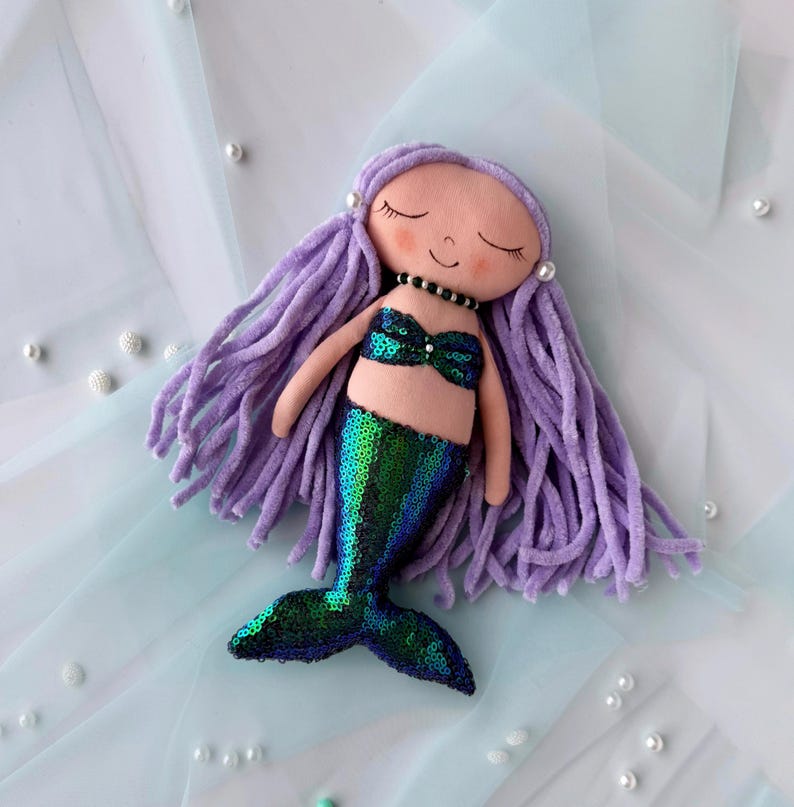 May include: A handmade mermaid doll with purple yarn hair, a green sequined tail, and a pink fabric top. The doll is lying on a light blue background.