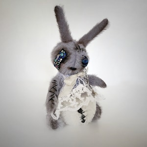 May include: A gray plush bunny rabbit with blue eyes and a white lace dress. The bunny has a jeweled eye patch and stitched details.