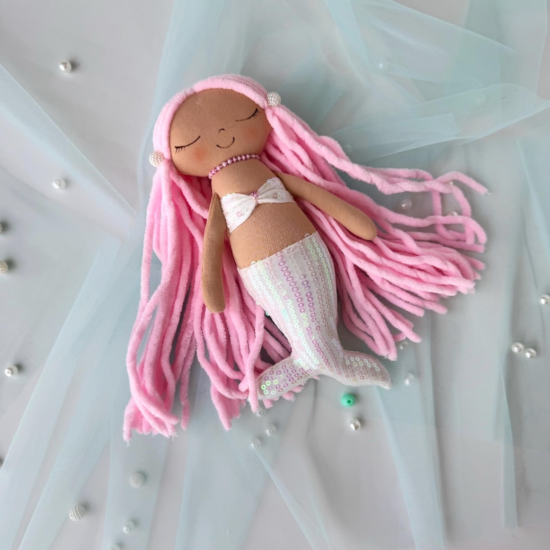 May include: A pink and white mermaid doll with long pink yarn hair and a white sequined tail. The doll is lying on a light blue tulle fabric.