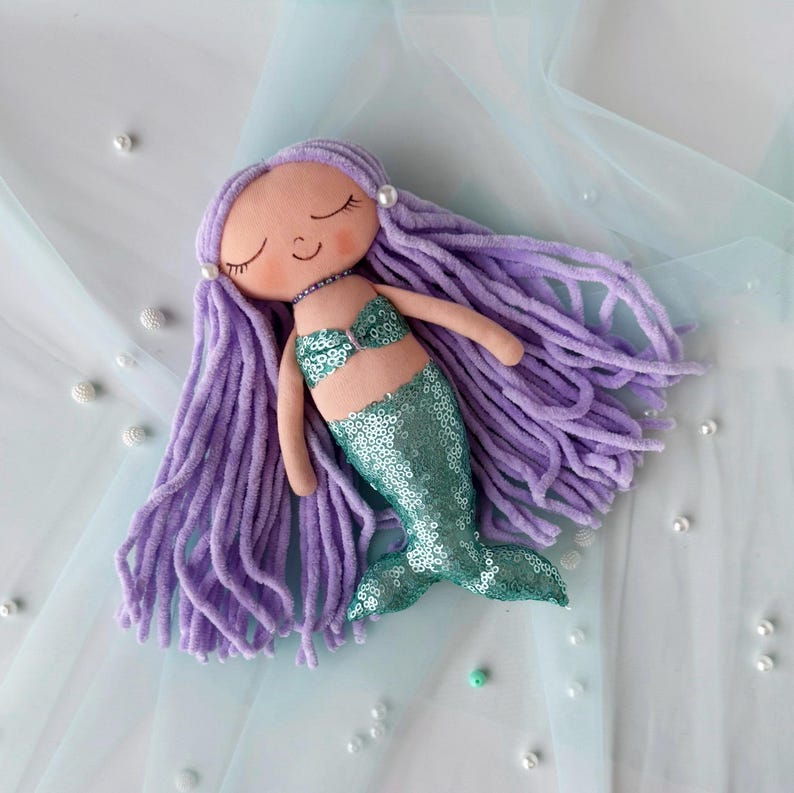 May include: A handmade purple haired mermaid doll with a green sequin tail. The doll has a smiling face with closed eyes and is wearing a light pink seashell bra.