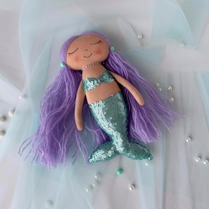 May include: A handmade mermaid doll with purple yarn hair, a teal sequin tail, and a light brown fabric bodice. The doll is lying on a light blue fabric background.