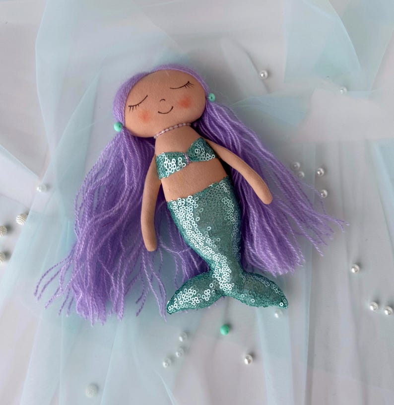 May include: A handmade mermaid doll with purple yarn hair, a teal sequin tail, and a light brown fabric bodice. The doll is lying on a light blue fabric background.