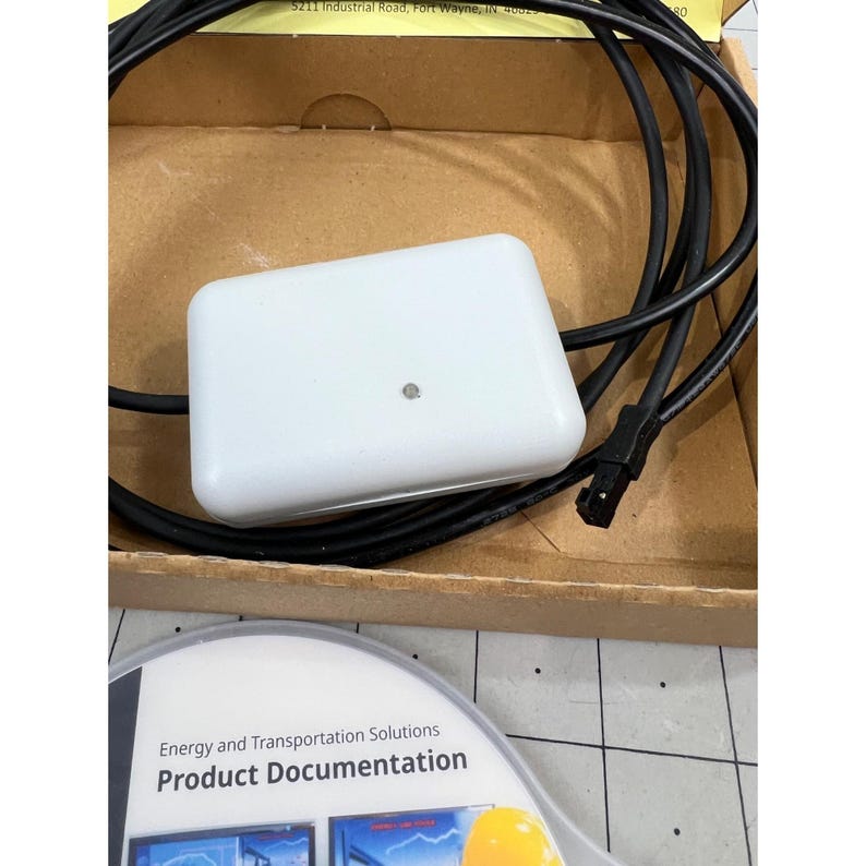 May include: A white rectangular electronic device with a black cable and plug, resting in a cardboard box. The device has a small, circular indicator light. Product documentation is visible below, with the text "Energy and Transportation Solutions".