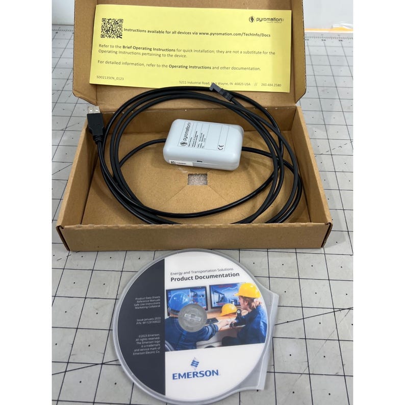 May include: An open cardboard box containing a white Pyromation device, a black cable, and a product documentation CD. The CD is in a clear plastic sleeve and features the Emerson logo.