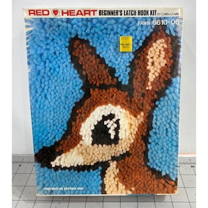May include: A Red Heart Beginner's Latch Hook Kit featuring a fawn design. The kit includes a blue background with a brown and white fawn image. The kit's title and brand name are displayed at the top. The kit's model number is 6610-06.