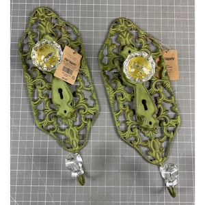 May include: Two decorative wall hooks with a distressed green finish. Each hook features a clear glass knob, a keyhole design, and a decorative filigree pattern. The hooks have a clear glass hook at the bottom and a tag that says "Pier 1 Imports".