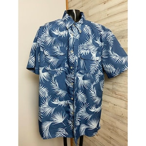 May include: Blue short-sleeved shirt featuring a white palm leaf design. The shirt has a button-down front, two chest pockets, and a collared neckline. The fabric has a subtle striped texture.