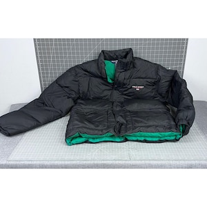 Könnte beinhalten: Schwarze Steppjacke mit grünem Innenfutter. Die Jacke hat einen hohen Kragen, zwei Vordertaschen und den Schriftzug "POLO SPORT" auf der Brust. Die Jacke liegt flach auf einer grauen Oberfläche.
