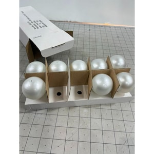 May include: A package of white, pearlescent Christmas ornaments. The ornaments are round and sit in individual cardboard dividers within a white box. The box is partially open, revealing the contents. The ornaments are a classic holiday decoration.