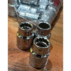 Puede incluir: Llaves de vaso cromadas brillantes con un diseño de 12 puntas. Las herramientas están dispuestas sobre una superficie de madera, algunas aún en una bolsa de plástico transparente. Estas llaves son para uso automotriz o mecánico.