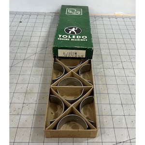 Peut inclure: Une boîte verte et marron de coussinets de moteur Toledo. La boîte est ouverte, révélant plusieurs coussinets de moteur de couleur argentée. La boîte porte le logo Toledo et le texte "ENGINE BEARINGS". La boîte est sur une surface grise.