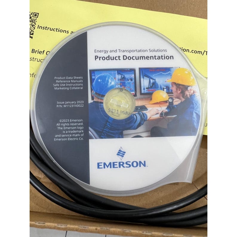 May include: A product documentation CD-ROM from Emerson, featuring the text "Energy and Transportation Solutions" and "Product Documentation." The disc includes images of people wearing hard hats and working on computers. The packaging also includes a black cable.