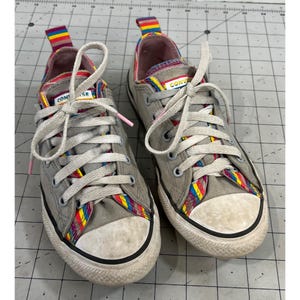 Op de afbeelding: Paar grijze Converse lage sneakers met witte veters en een witte neus. De sneakers hebben kleurrijke regenboogaccenten op de tong en rond de neus. Het woord "CONVERSE" is zichtbaar op de tong.