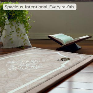 May include: A beige prayer rug with a white floral design and a white border. An open book on a wooden stand is in the background. The text "Spacious. Intentional. Every rak'ah." is at the top.