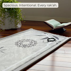 Op de afbeelding: Een lichtgrijs gebedskleed met een centraal geometrisch ontwerp en bloemaccenten. Het kleed is omrand in een donkerder grijs en heeft de tekst "Spacious. Intentional. Every rak'ah." Een gebedsketting en een open boek zijn ook zichtbaar.