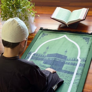 May include: A person wearing a white cap and dark clothing is kneeling on a green prayer rug with a depiction of the Kaaba. An open book rests on a wooden stand, and a potted plant is in the background. The scene is set on a wooden floor.