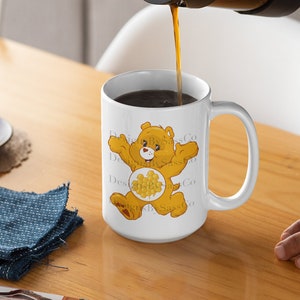 Peut inclure: Une tasse en céramique blanche avec un personnage de dessin animé Care Bear, jaune avec un symbole de nid d'abeilles sur le ventre. Du café est versé dans la tasse à partir d'une cafetière à piston. Un petit gâteau et un tissu bleu sont sur la table en bois.
