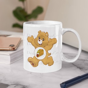Pode incluir: Caneca de cer&acirc;mica branca com um urso de desenho animado com uma taco na barriga. O urso &eacute; marrom claro com um rosto sorridente e bra&ccedil;os estendidos. A caneca tem uma al&ccedil;a curva e o texto "Designs By Sass Co" est&aacute; impresso.