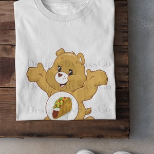 Pode incluir: Camiseta branca com um urso de desenho animado e um gr&aacute;fico de taco. O urso &eacute; marrom claro com um cora&ccedil;&atilde;o no peito. O taco tem v&aacute;rios recheios. A camiseta est&aacute; em uma superf&iacute;cie de madeira, com t&ecirc;nis brancos na parte inferior.