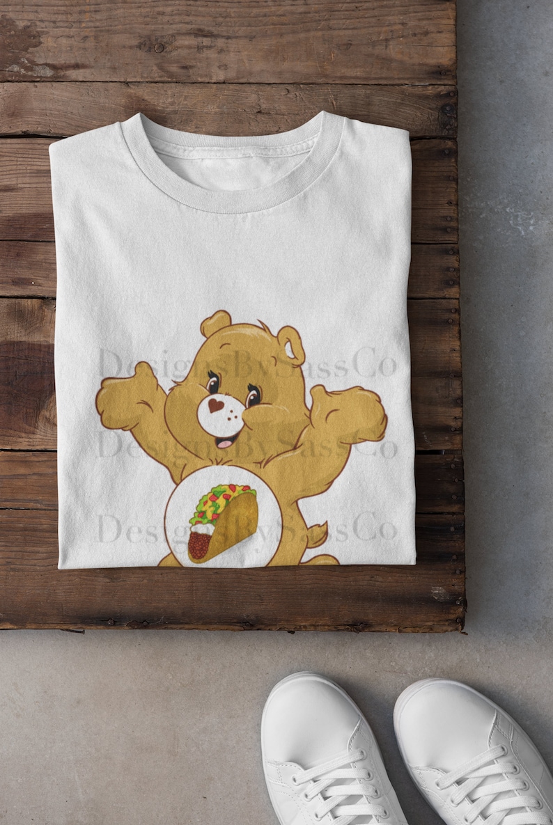 Pode incluir: Camiseta branca com um urso de desenho animado e um gr&aacute;fico de taco. O urso &eacute; marrom claro com um cora&ccedil;&atilde;o no peito. O taco tem v&aacute;rios recheios. A camiseta est&aacute; em uma superf&iacute;cie de madeira, com t&ecirc;nis brancos na parte inferior.