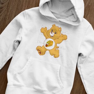 Pode incluir: Moletom branco com um urso de desenho animado e um gr&aacute;fico de taco. O urso &eacute; marrom claro com um cora&ccedil;&atilde;o vermelho no p&eacute;. O moletom tem um bolso canguru e um capuz.