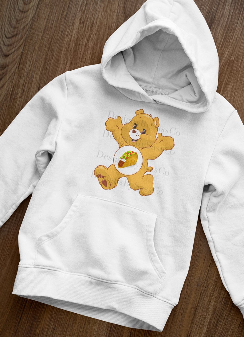Pode incluir: Moletom branco com um urso de desenho animado e um gr&aacute;fico de taco. O urso &eacute; marrom claro com um cora&ccedil;&atilde;o vermelho no p&eacute;. O moletom tem um bolso canguru e um capuz.
