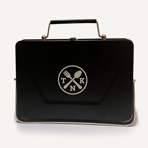Puede incluir: Fiambrera de metal negro con asa plateada y un logotipo plateado de un tenedor y una cuchara cruzados con las letras "TKN" en un c&iacute;rculo.