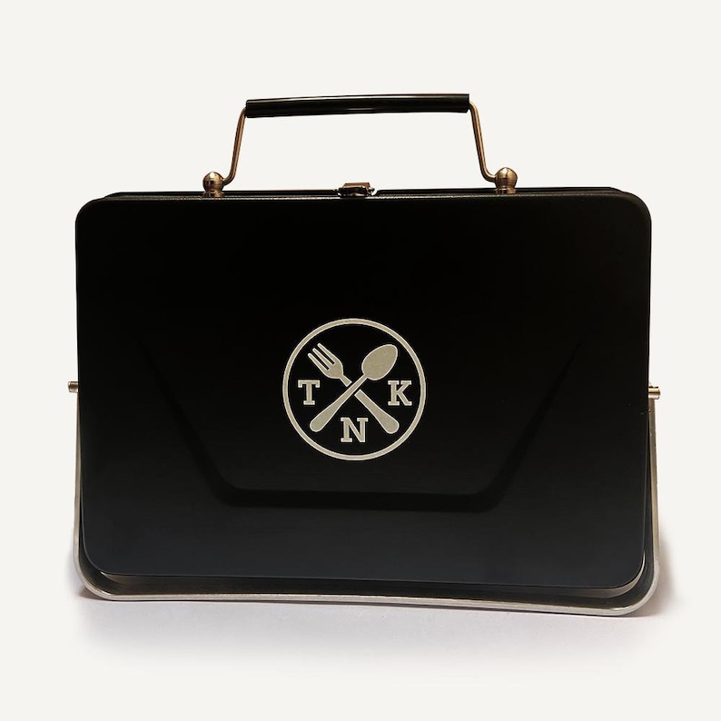 Puede incluir: Fiambrera de metal negro con asa plateada y un logotipo plateado de un tenedor y una cuchara cruzados con las letras "TKN" en un c&iacute;rculo.