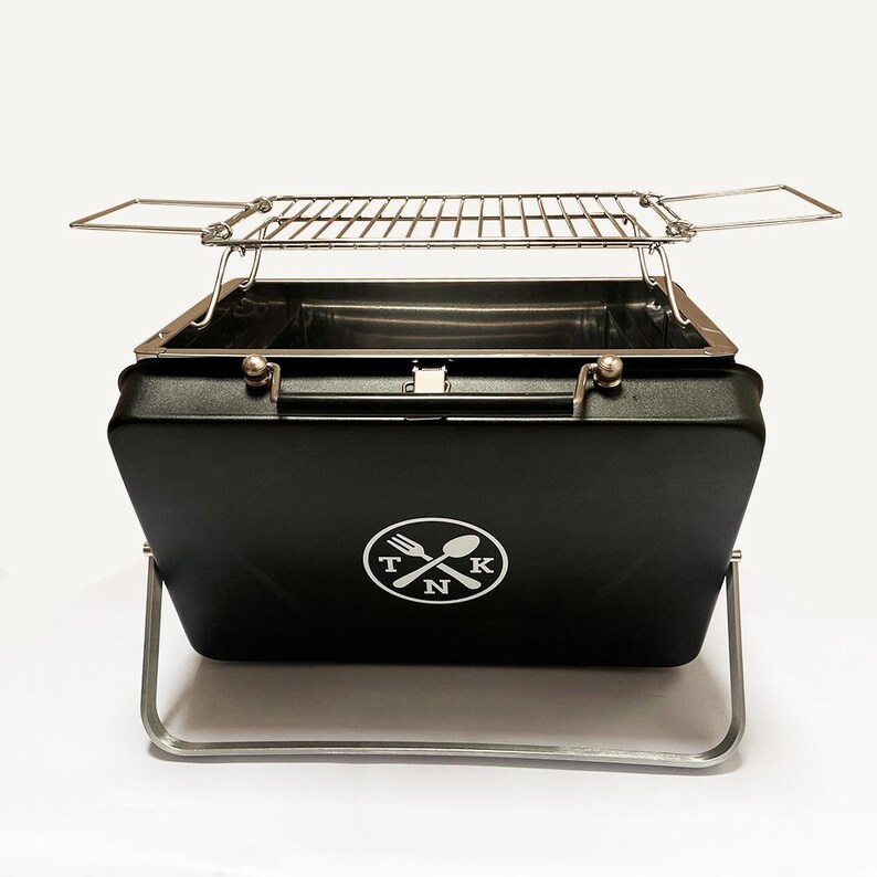 Puede incluir: Una barbacoa de carb&oacute;n port&aacute;til negra con una rejilla de cocci&oacute;n cromada y un logotipo que dice "TNK" con un tenedor y una cuchara cruzados dentro de un c&iacute;rculo. La barbacoa tiene un asa y patas plegables.