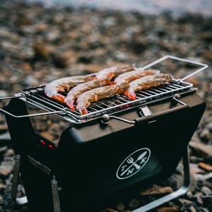 Puede incluir: Una parrilla port&aacute;til negra con un asa plateada y un logotipo de un tenedor y una cuchara cruzados con las letras "TNK". La parrilla est&aacute; sobre un lecho de peque&ntilde;as piedras y tiene camarones cocin&aacute;ndose en la parrilla.