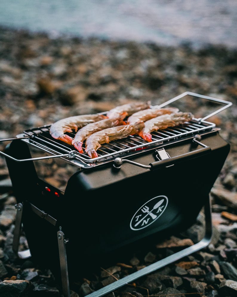 Puede incluir: Una parrilla port&aacute;til negra con un asa plateada y un logotipo de un tenedor y una cuchara cruzados con las letras "TNK". La parrilla est&aacute; sobre un lecho de peque&ntilde;as piedras y tiene camarones cocin&aacute;ndose en la parrilla.