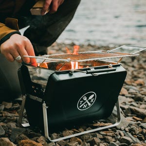 Puede incluir: Una parrilla port&aacute;til negra con una rejilla plateada y un logotipo blanco que dice "TNK" con un tenedor y una cuchara. La parrilla est&aacute; instalada en una superficie rocosa con llamas visibles en el interior.