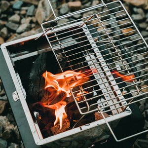 Puede incluir: Una peque&ntilde;a parrilla de carb&oacute;n port&aacute;til con una rejilla de metal y un fuego ardiendo en su interior. La parrilla est&aacute; hecha de acero inoxidable y tiene un acabado negro.