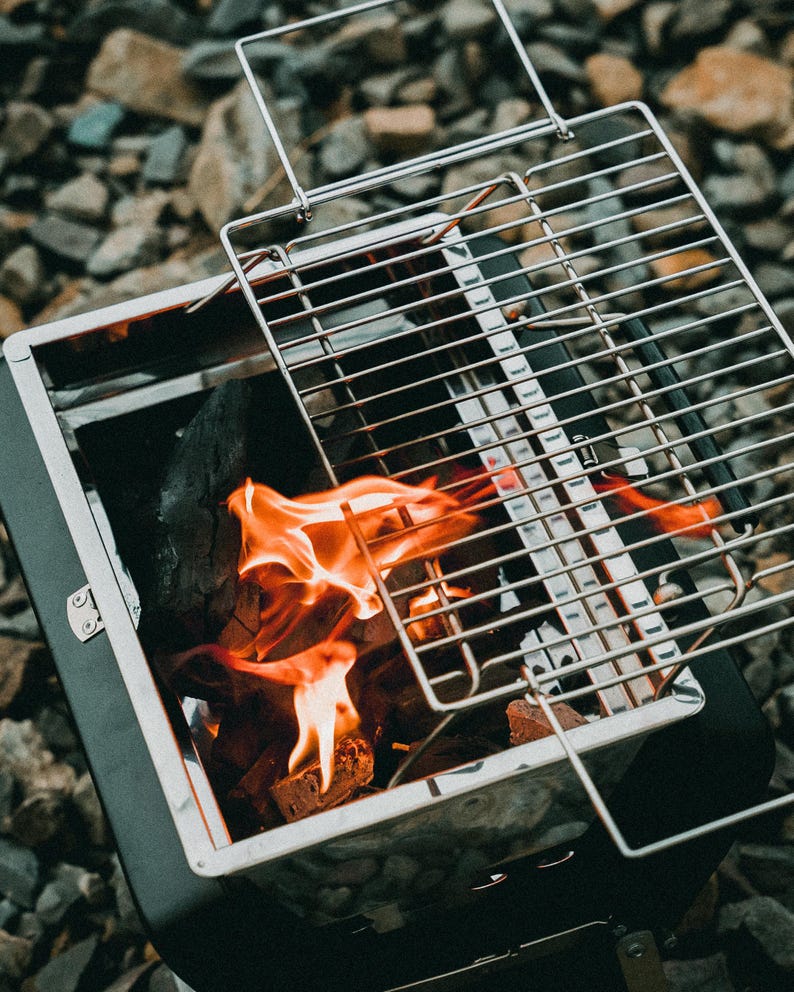 Puede incluir: Una peque&ntilde;a parrilla de carb&oacute;n port&aacute;til con una rejilla de metal y un fuego ardiendo en su interior. La parrilla est&aacute; hecha de acero inoxidable y tiene un acabado negro.