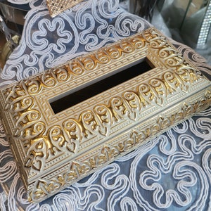 Op de afbeelding: Gouden tissuebox met een versierd ontwerp. De doos heeft een witte achtergrond met gouden accenten en ingewikkelde patronen.