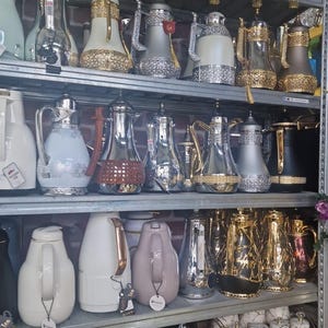 May include: A display of various vacuum insulated thermal carafes and coffee pots arranged on metal shelving. The collection features diverse finishes including polished chrome, matte white, black, gold, and silver accents with ornate filigree details.
