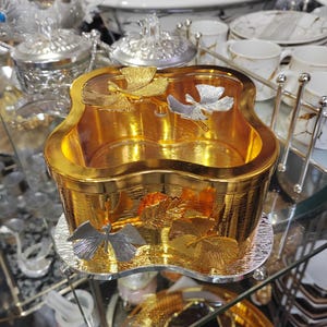 May include: A gold-colored decorative bowl with a unique shape, featuring silver and gold leaf accents. The bowl sits on a mirrored surface, reflecting the intricate details of the design. Other tableware items are visible in the background.