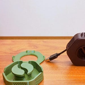 May include: A brown hexagonal cable organizer with a USB connector and a smaller connector. A green, two-part cable holder is nearby. A bronze Buddha statue sits in the background on a wooden surface.