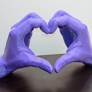 May include: A pair of purple hands forming a heart shape. The hands are positioned against a neutral background, with the fingers of each hand touching to create the heart symbol. The hands are a solid, smooth color.