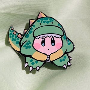 Puede incluir: Un pin de esmalte verde y amarillo de un personaje rosa que lleva un disfraz de dinosaurio verde. El personaje tiene ojos grandes y una boca pequeña.