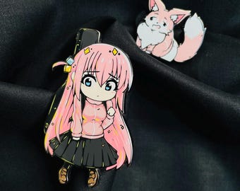 Puede incluir: Dos pines de esmalte con personajes de anime. El primer pin es de una chica con cabello rosado, usando un suéter rosado y una falda negra. El segundo pin es de un zorro rosado con pelaje blanco.